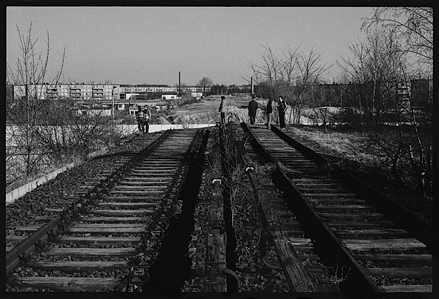 Photographie noir et blanc de rails de train coupé plus loin par le mur, quelques personnes discutent sur les rails