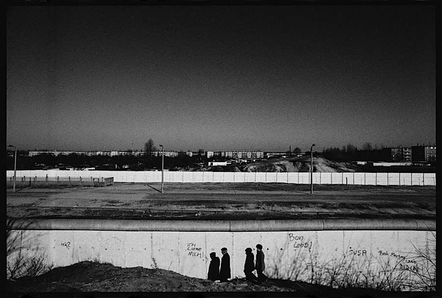 Photographie noir et blanc éloignée du mur ainsi que du no man's land, deux personnes marchent tranquillement aux abords du mur